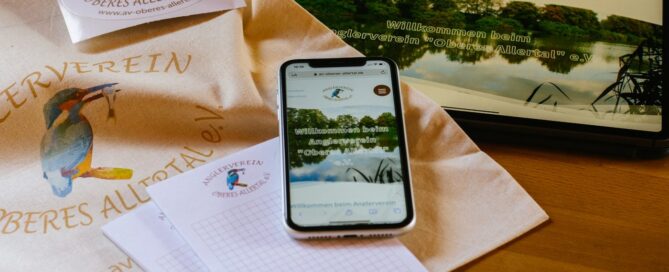 Website des Anglervereins auf dem Handy, dazu Tasche und Aufkleber mit Logo